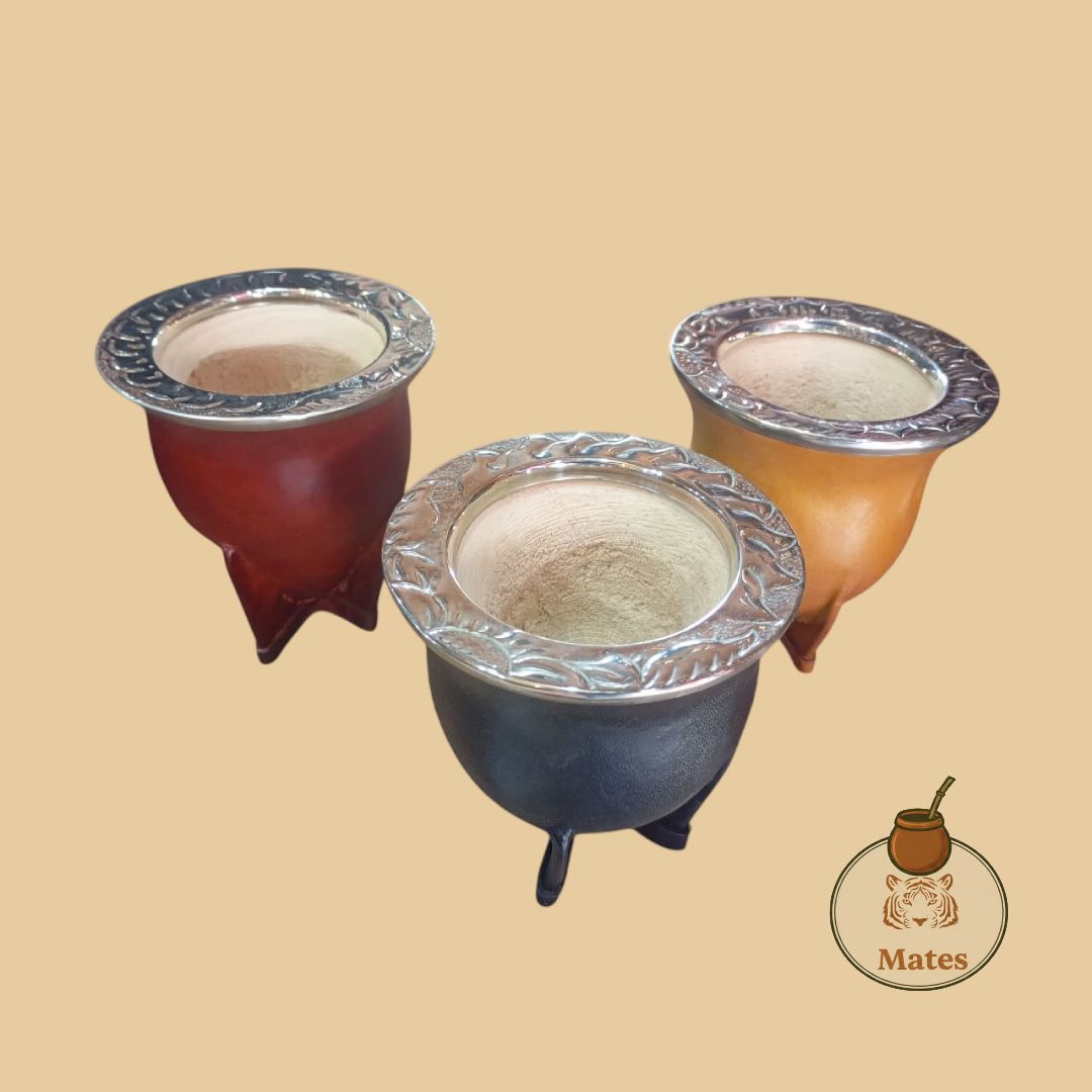 Tres calabazas de yerba mate en rojo, marrón y negro con bordes y patas plateadas, sobre un fondo marrón.