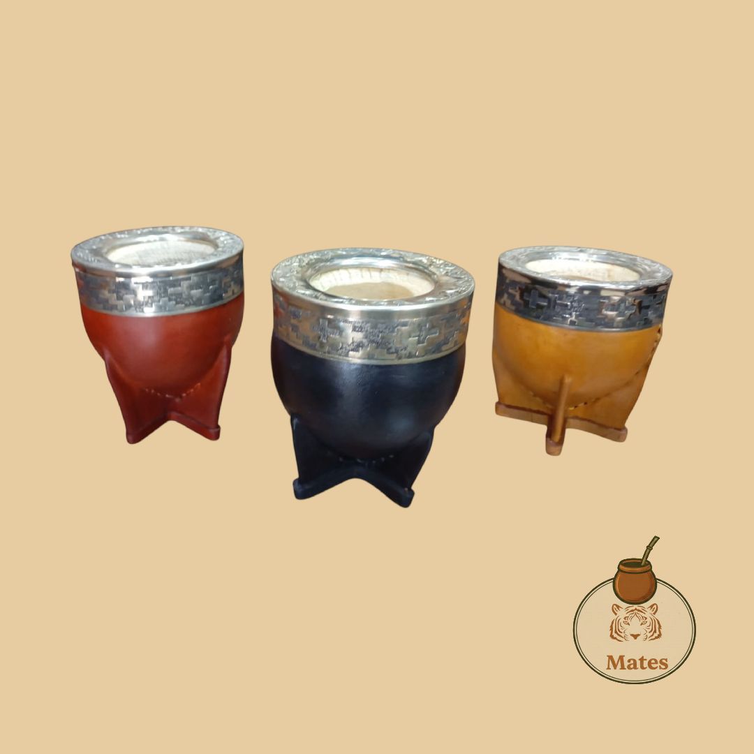 Tres tazas de mate, roja, negra y amarilla, con bordes plateados y soportes de madera, sobre un fondo tostado.