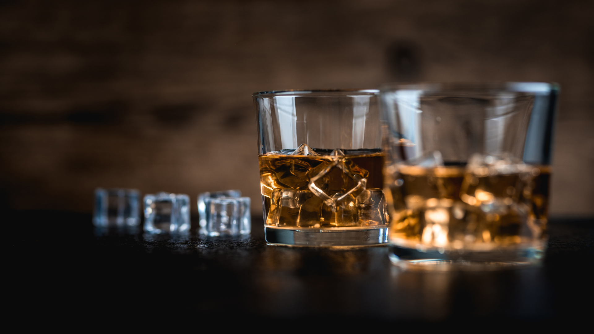 Three beers in glasses on a wooden table, with kegs stacked in the background under a blue sky.