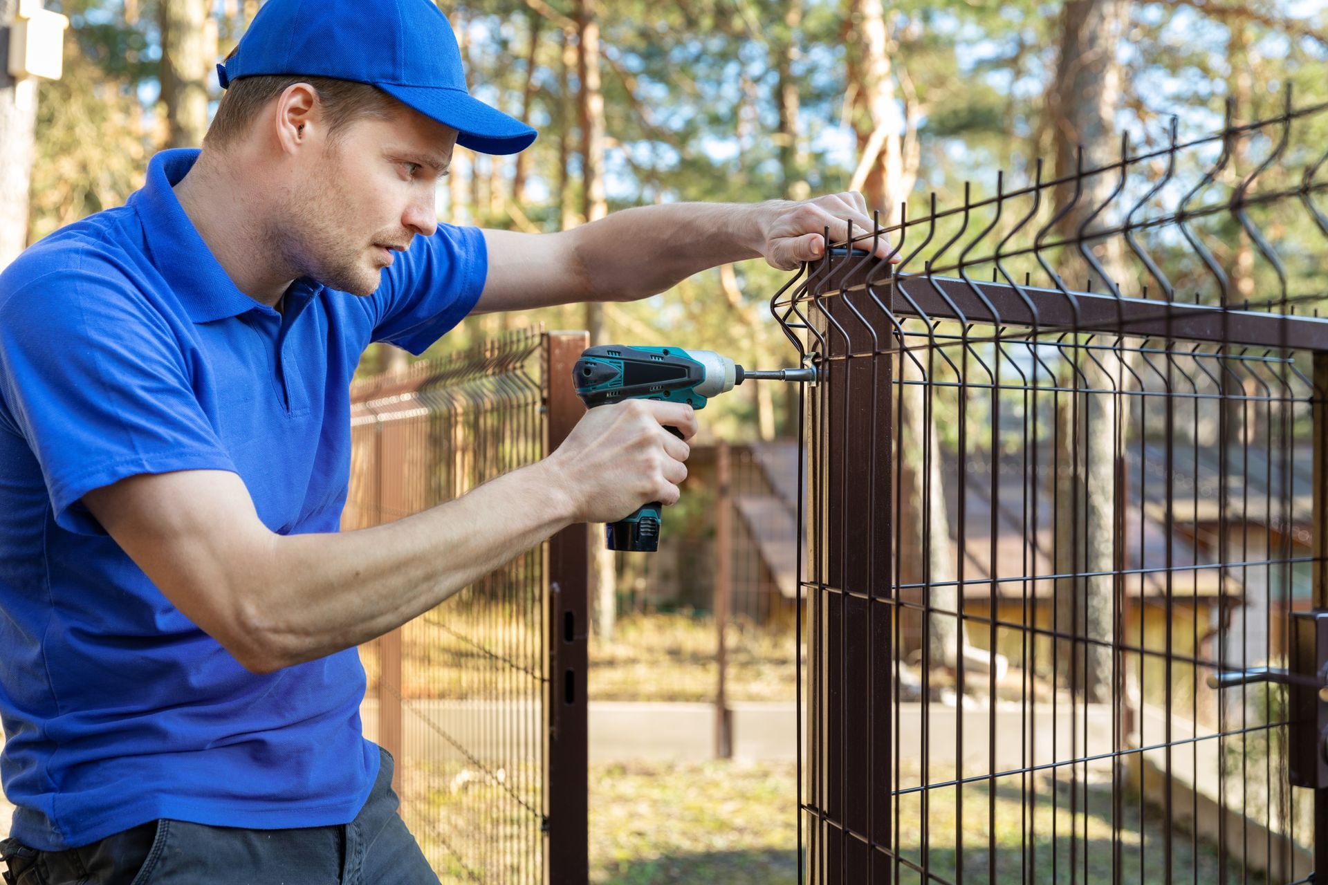 Man in blue shirt and cap uses a power drill to install a brown metal fence outdoors.