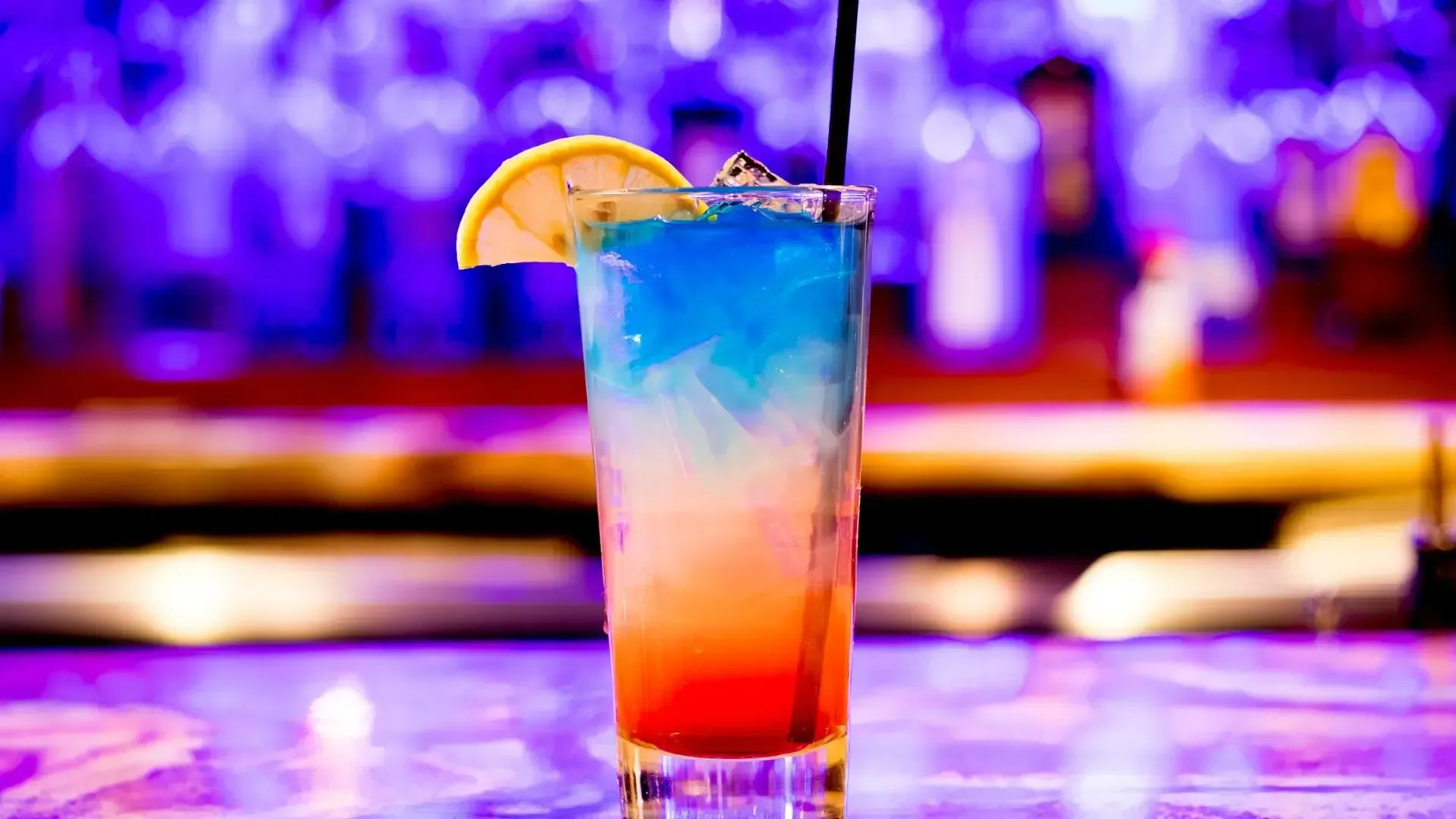 A close up of a colorful cocktail with a straw on a bar.