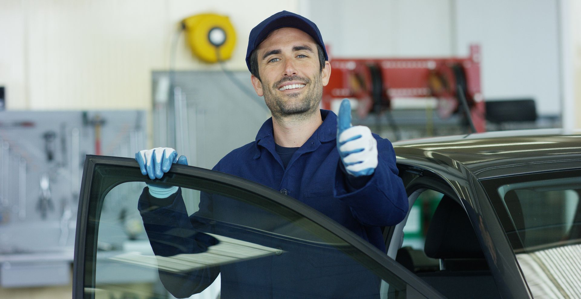 Auto mechanic in blue uniform holding car window, giving thumbs up in garage.