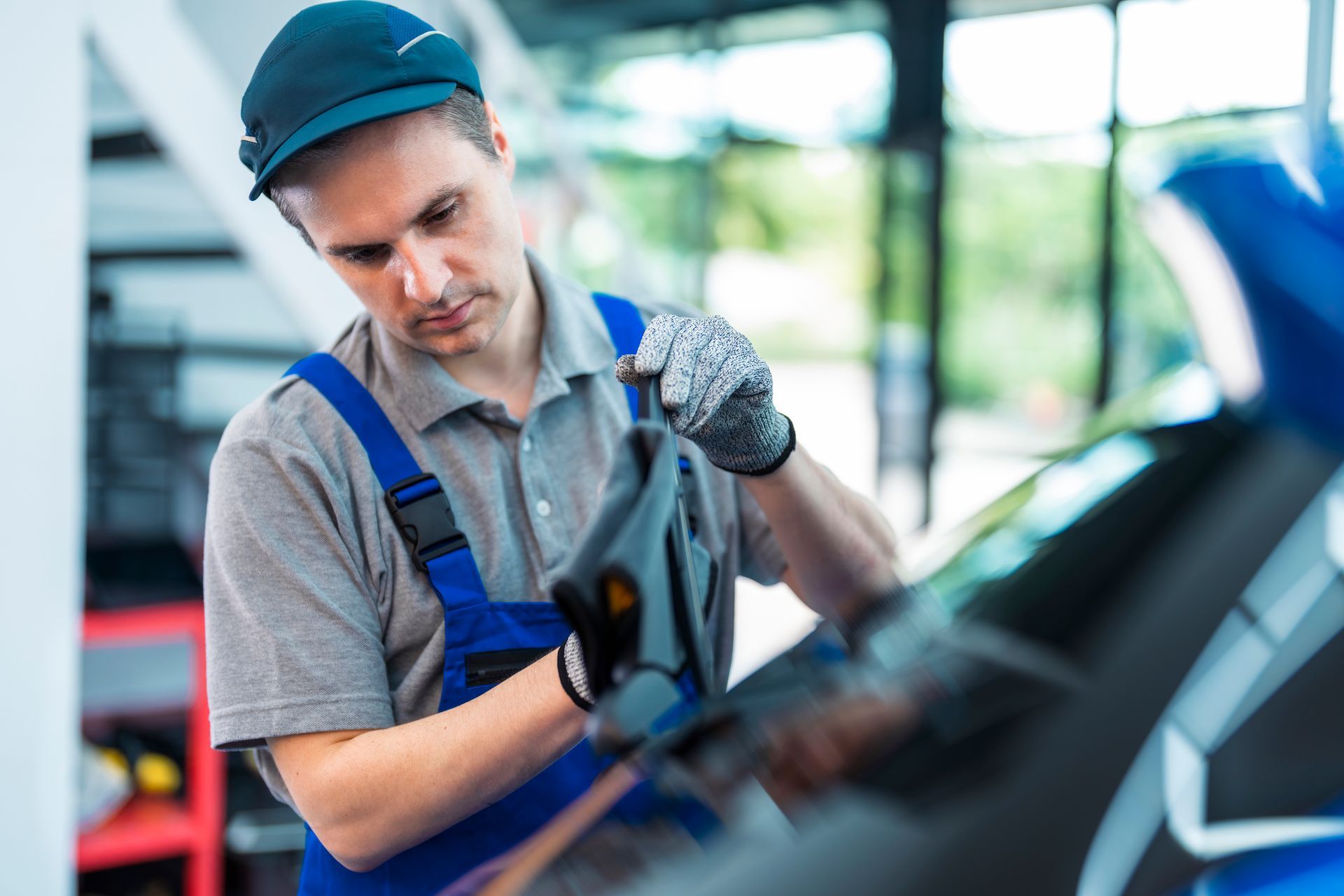 Mechanic in blue overalls fixing a car windshield indoors, wearing a cap and gloves.