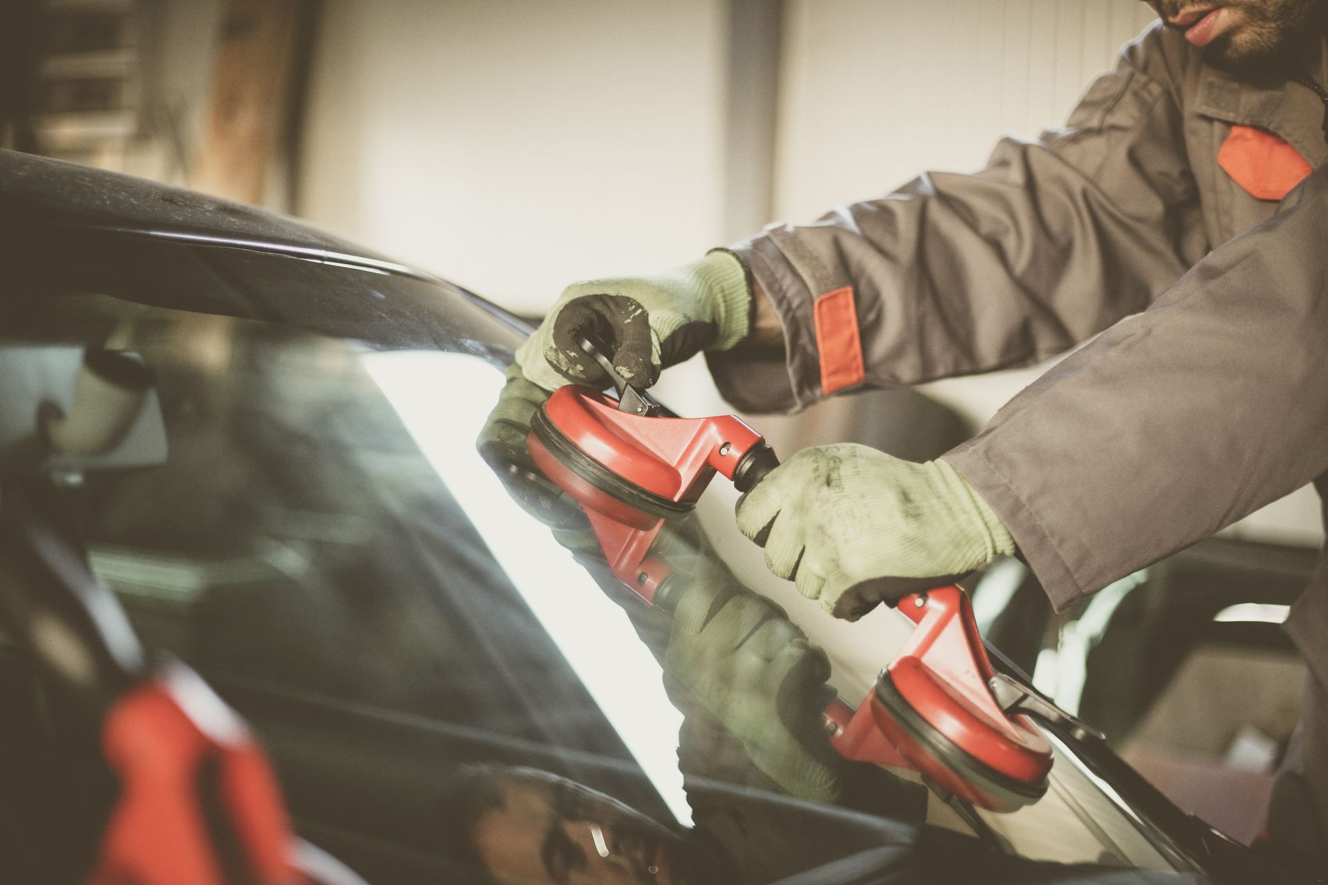 Mechanic in overalls using a suction cup tool to remove a windshield from a car.