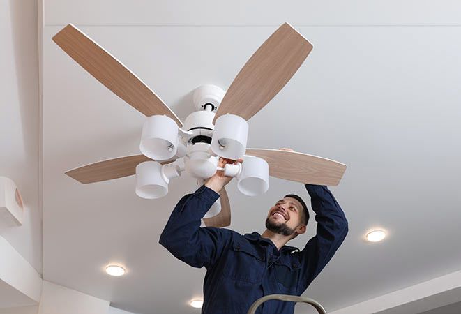 Man on a ladder changing a lightbulb in a ceiling fan, smiling. Indoors, white ceiling.