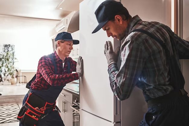 Two men in work clothes push a white refrigerator in a kitchen.