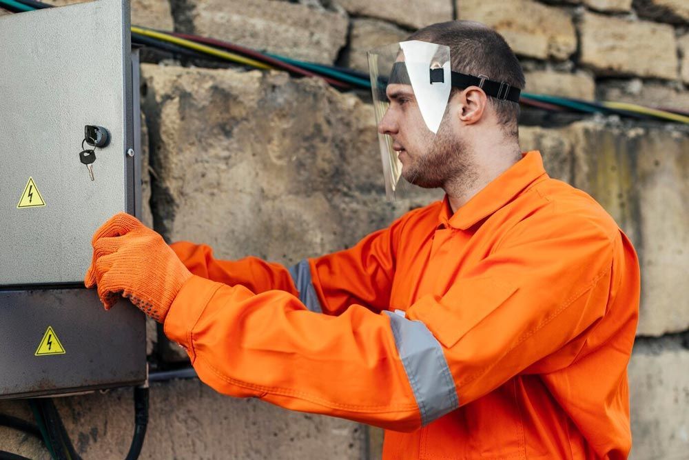 Man in orange work suit, face shield, and gloves working on electrical box outdoors.