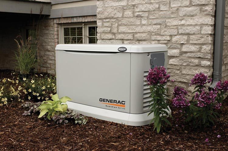 Gray Generac home backup generator next to a brick wall and flowers in an outdoor setting.