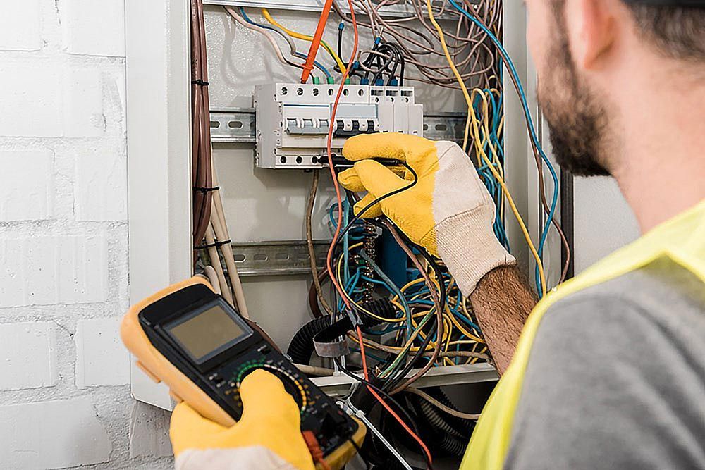 Electrician in yellow gloves using multimeter on a circuit breaker panel.