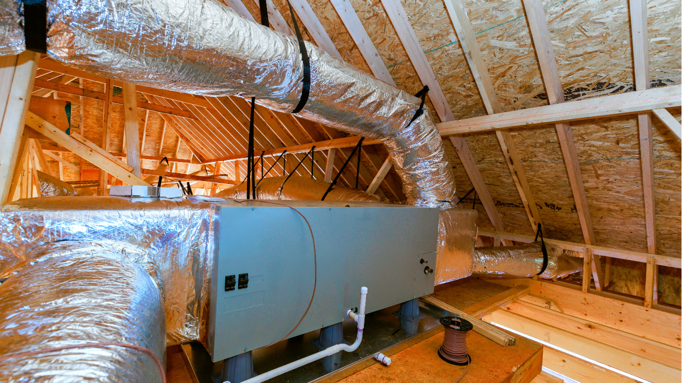 A gray HVAC air handler unit with silver flexible ductwork installed in an unfinished wooden attic.