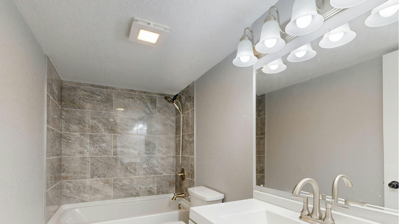 A modern bathroom with a grey tiled shower, a vanity mirror with light fixtures, and a white sink.