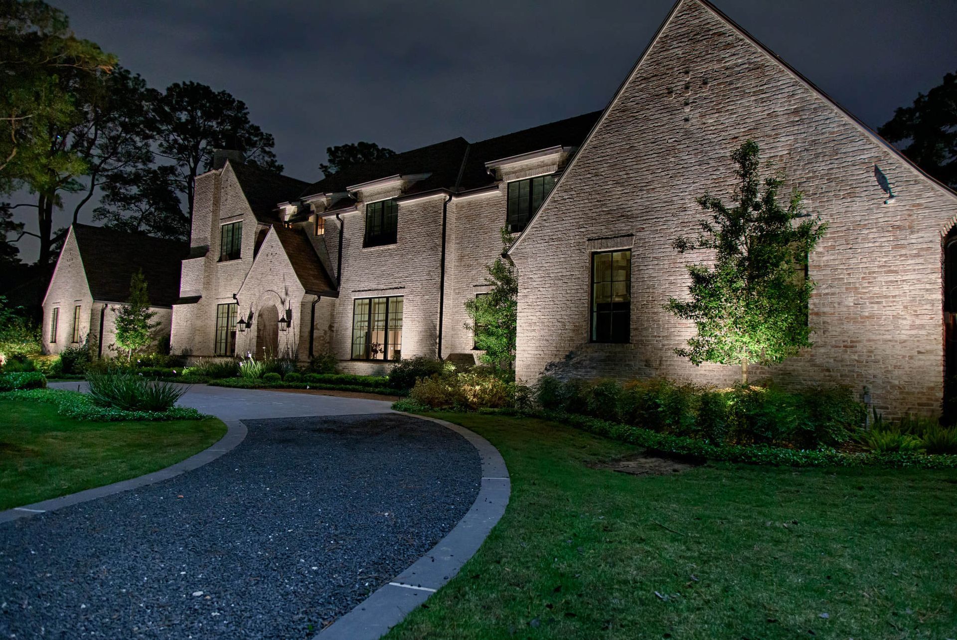 Night-lit brick house with a winding driveway, trees, and green grass.