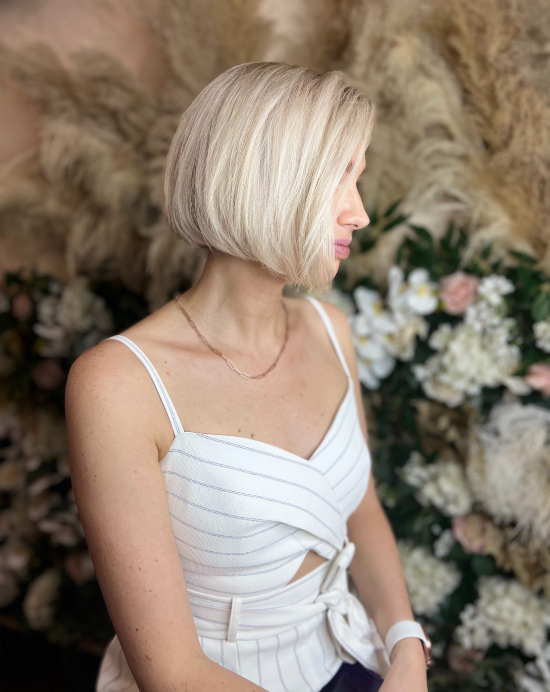 A woman in a white tank top is sitting in front of a wall of flowers.