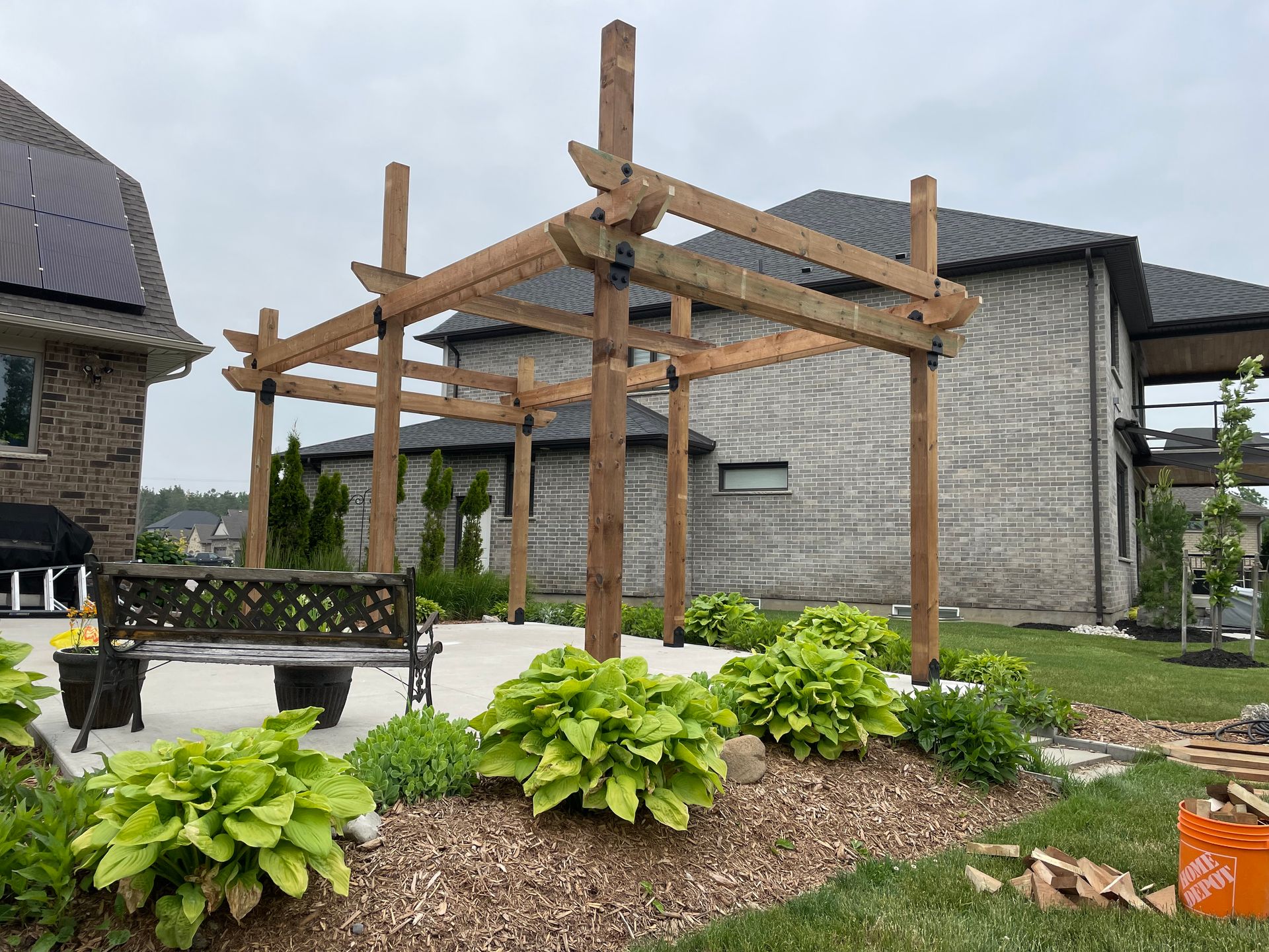 A wooden pergola is sitting in the middle of a garden in front of a house.