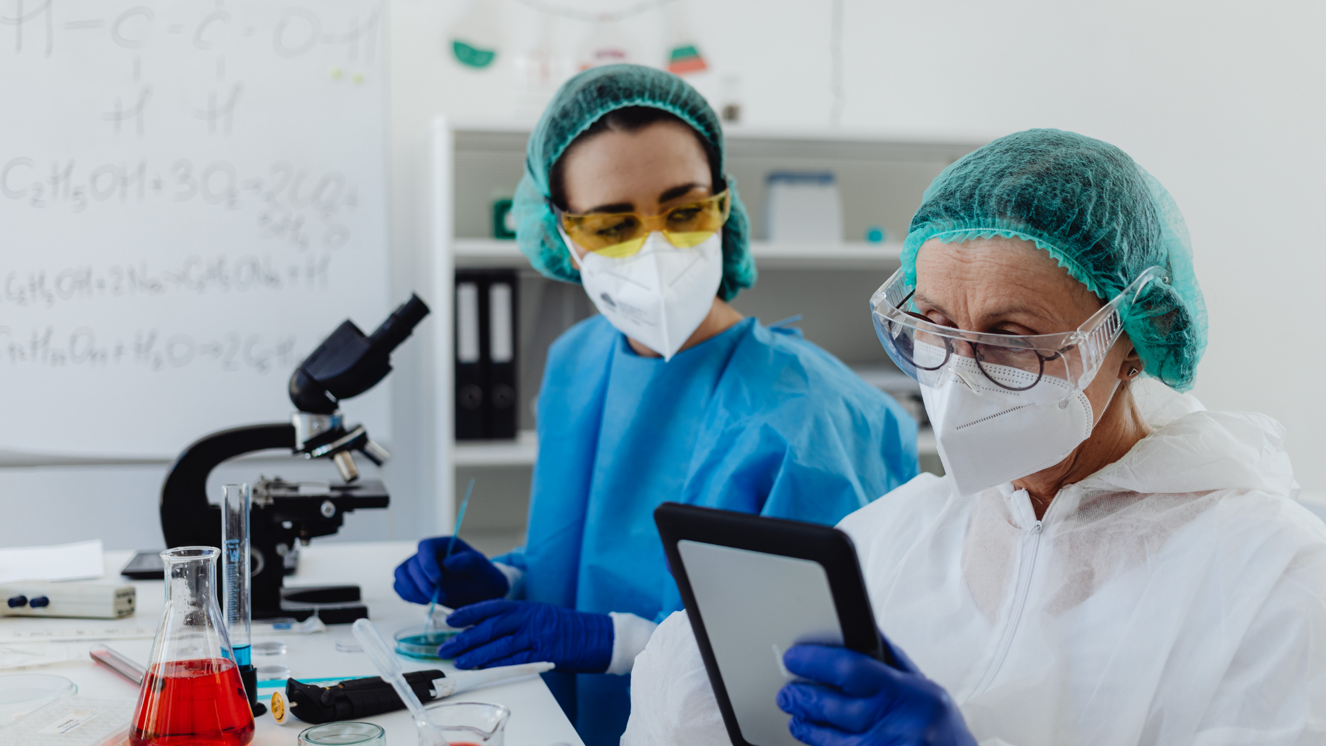 Two lab technicians in masks and gloves working with a microscope and tablet in a laboratory