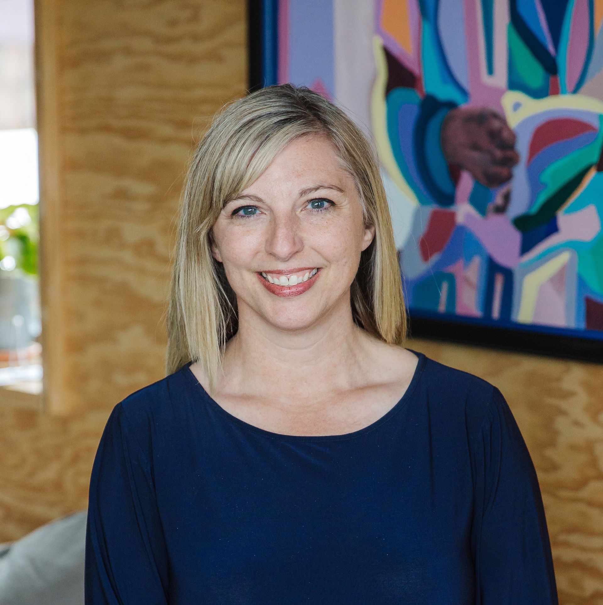 Kim Zacher smiling and sitting on the arm of a gray couch. She is inside a bright room, wearing a dark blue long-sleeved dress with teal on the sleeves and bottom of the dress. In the background, there is a window to the left with a small green plant on the window sill, a tan-colored wooden wall, with a colorful abstract picture with a black border.