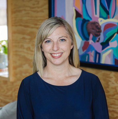 Kim Zacher smiling and sitting on the arm of a gray couch. She is inside a bright room, wearing a dark blue long-sleeved dress with teal on the sleeves and bottom of the dress. In the background, there is a window to the left with a small green plant on the window sill, a tan-colored wooden wall, with a colorful abstract picture with a black border.