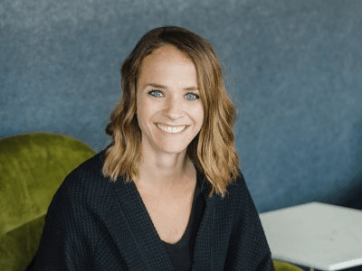Woman with wavy brown hair smiles, sitting on green chair in front of a blue wall.