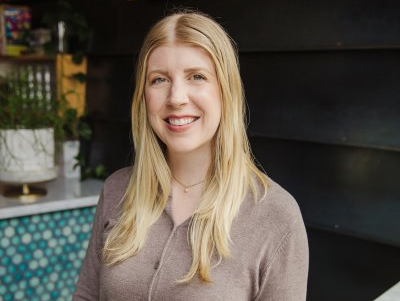 Woman with blonde hair smiles outside. Brown sweater, teal and white tiled counter visible.