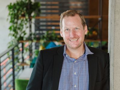 Man in a blazer and blue shirt smiles, leaning against a concrete column in a modern setting.