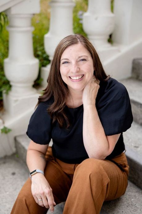 Kim Zacher smiling and sitting on the arm of a gray couch. She is inside a bright room, wearing a dark blue long-sleeved dress with teal on the sleeves and bottom of the dress. In the background, there is a window to the left with a small green plant on the window sill, a tan-colored wooden wall, with a colorful abstract picture with a black border.