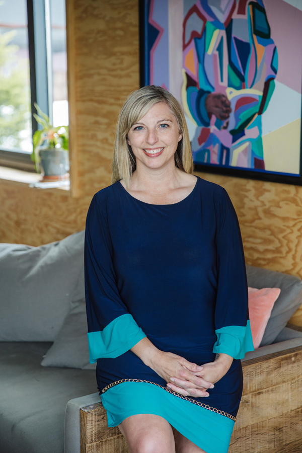 Kim Zacher smiling and sitting on the arm of a gray couch. She is inside a bright room, wearing a dark blue long-sleeved dress with teal on the sleeves and bottom of the dress. In the background, there is a window to the left with a small green plant on the window sill, a tan-colored wooden wall, with a colorful abstract picture with a black border.