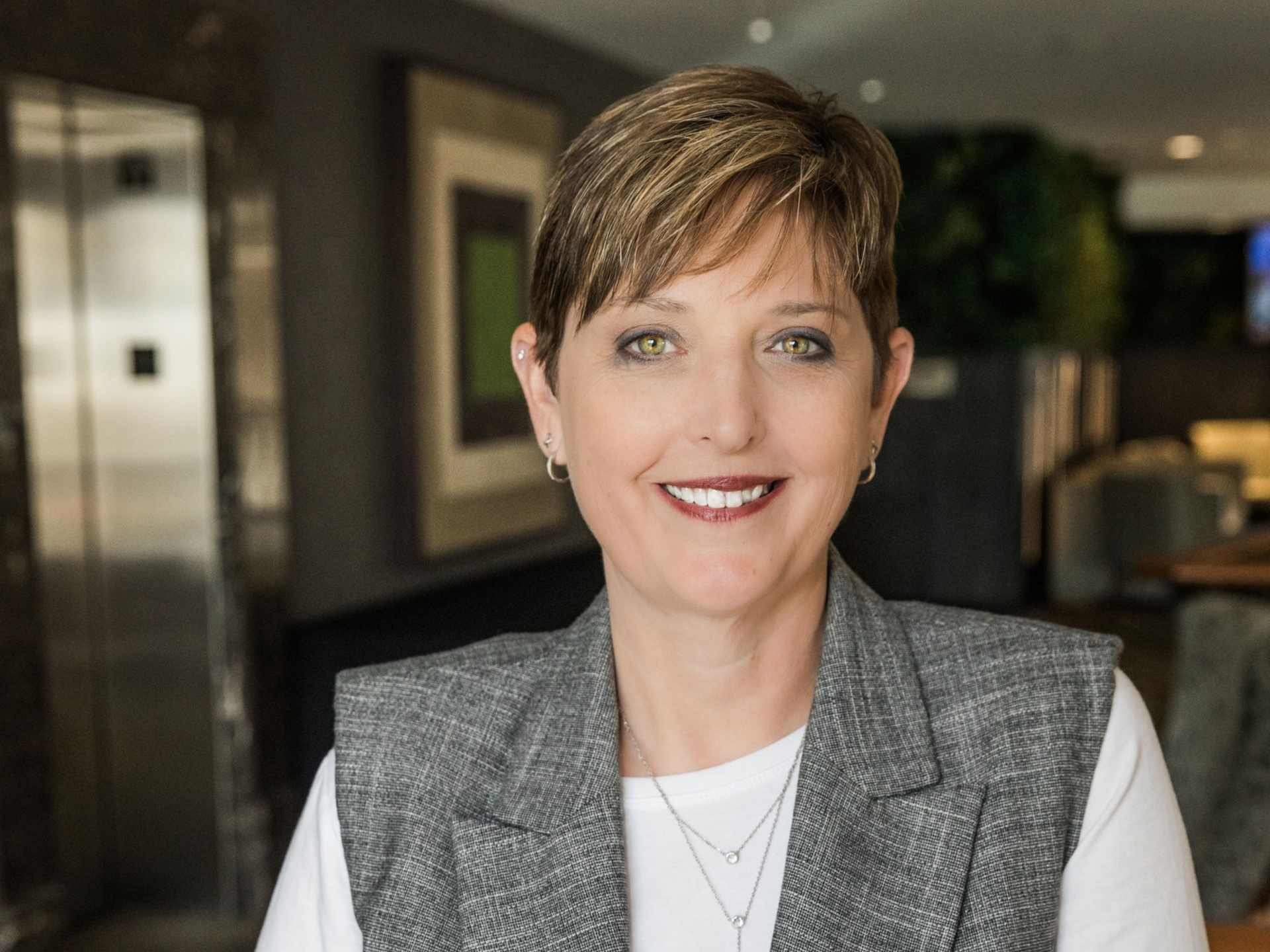 Woman in gray vest smiles, in a hotel lobby setting, near an elevator.