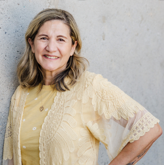 Woman in yellow top and lace cardigan smiles, leaning against a gray wall.