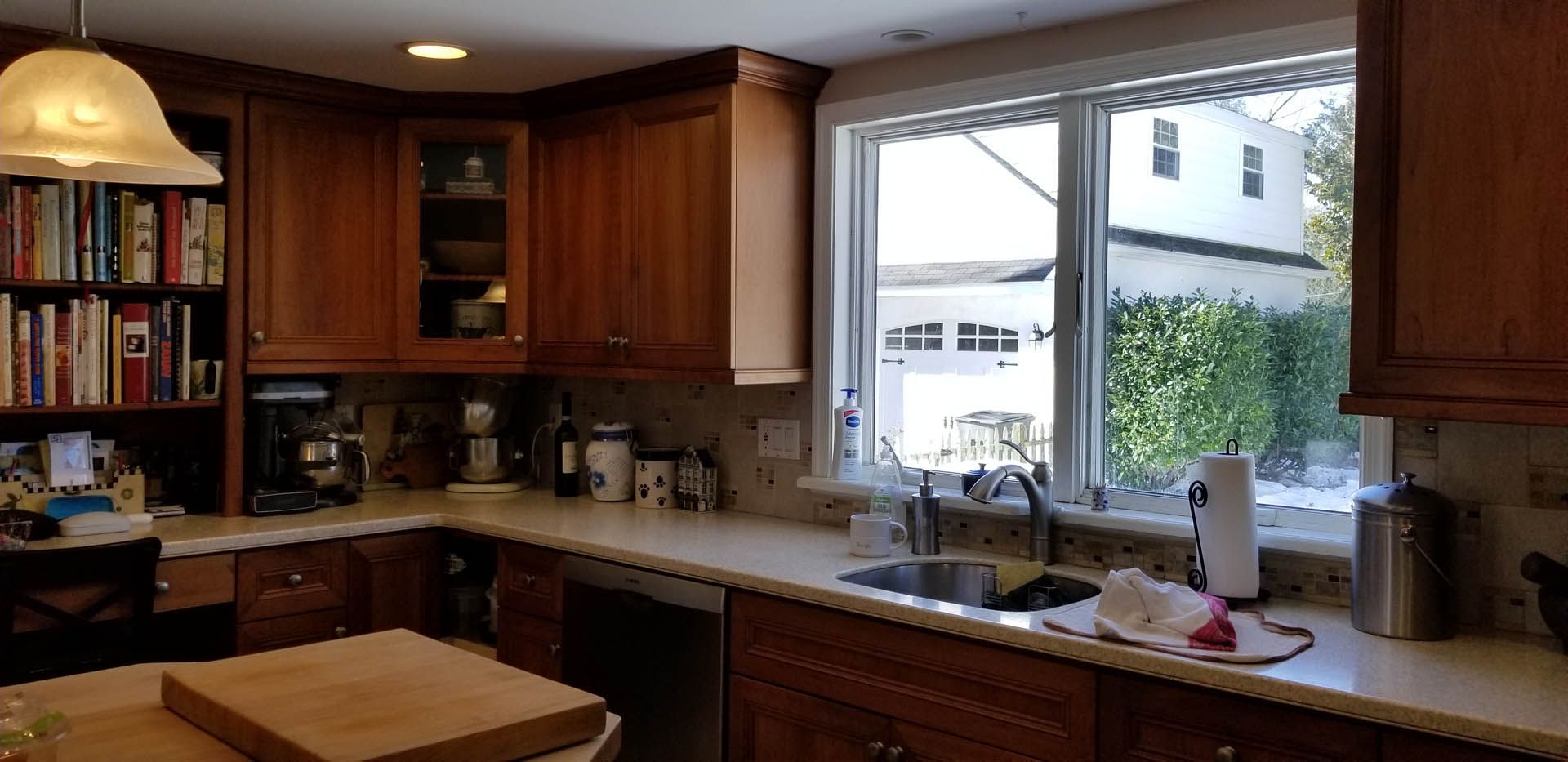 A kitchen with wooden cabinets, a countertop, a sink by a window, and a built-in bookshelf filled with books.