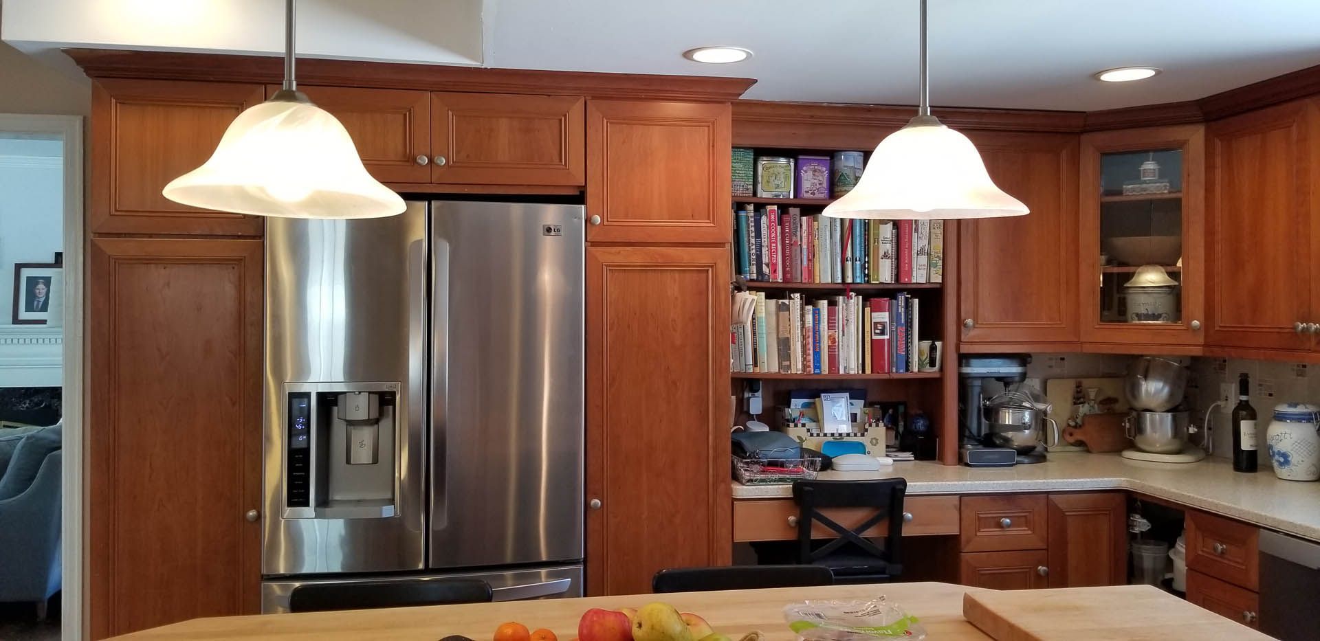 A kitchen with wooden cabinets, a stainless steel refrigerator, an open bookshelf, and hanging pendant lights.