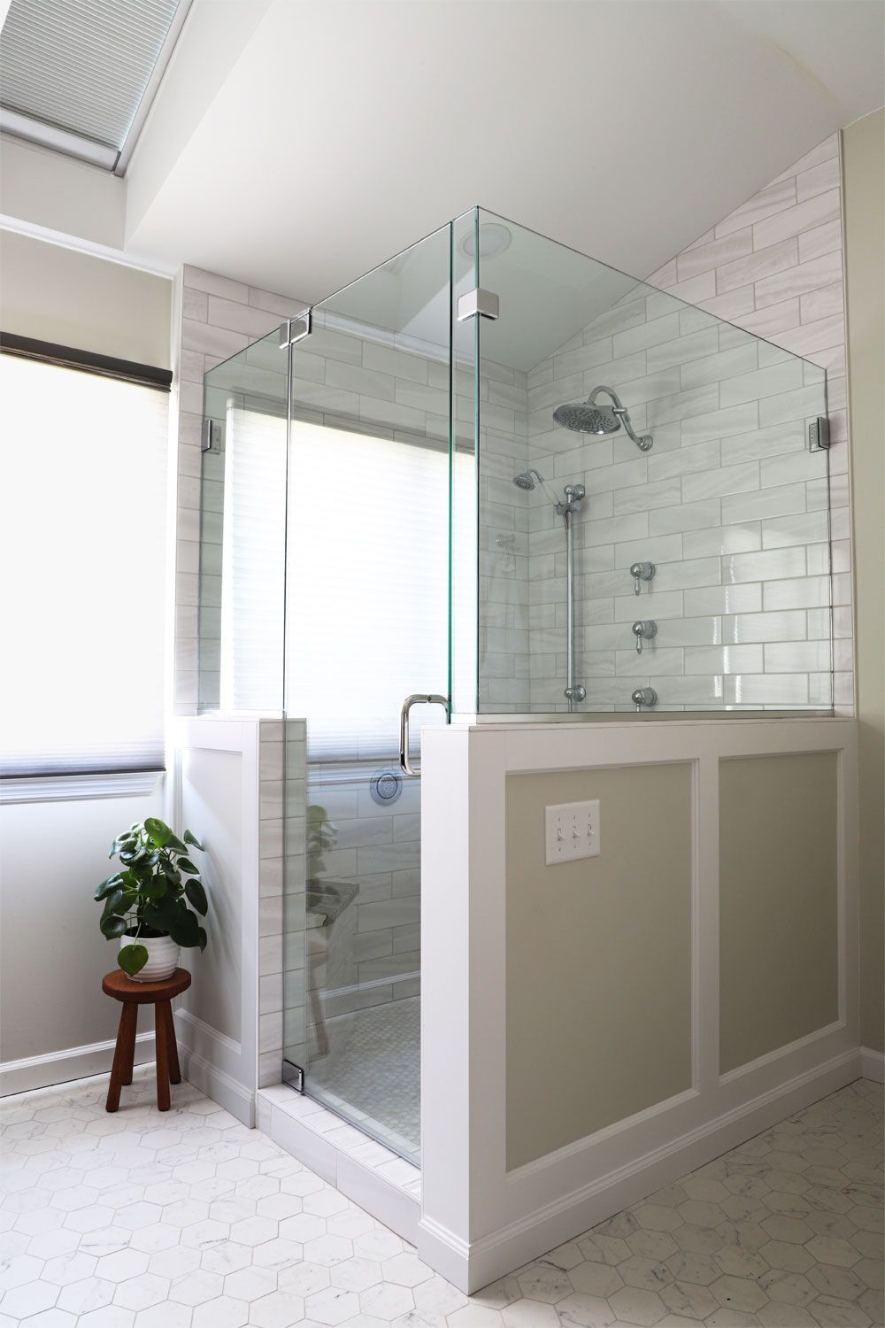 Glass shower enclosure with white wainscoting and subway tile. A small stool with a plant sits next to the window.