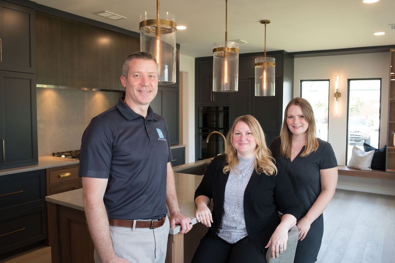 Three people pose in KBE Design Build's showroom with dark cabinetry and island; one man, two women.