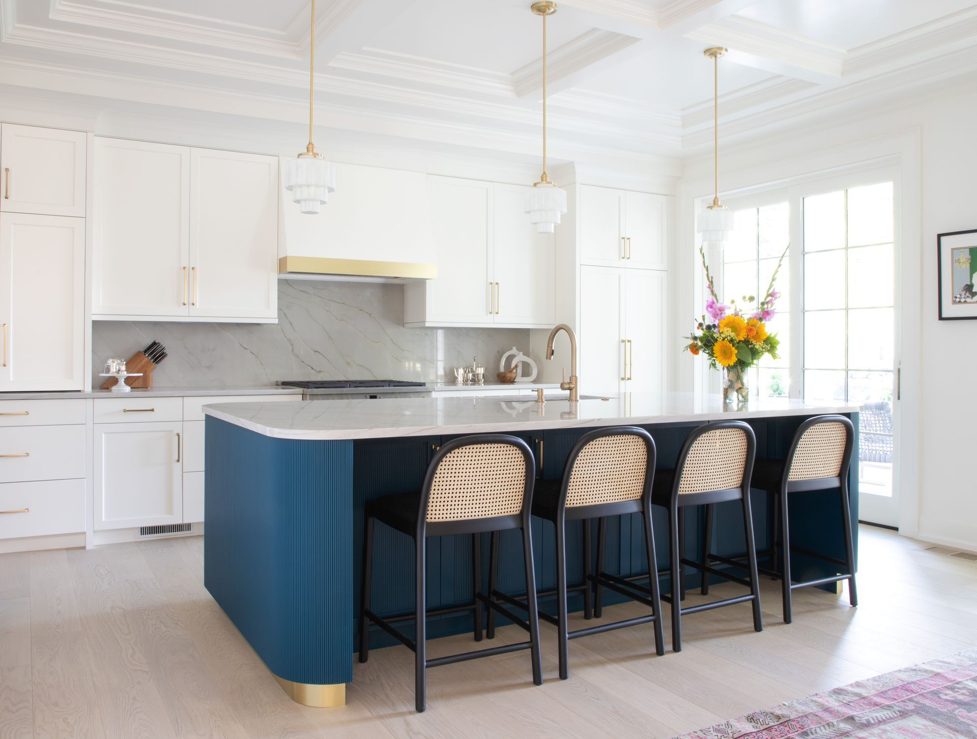 Blue and white kitchen with island, four rattan bar stools, gold accents, and flower arrangement.