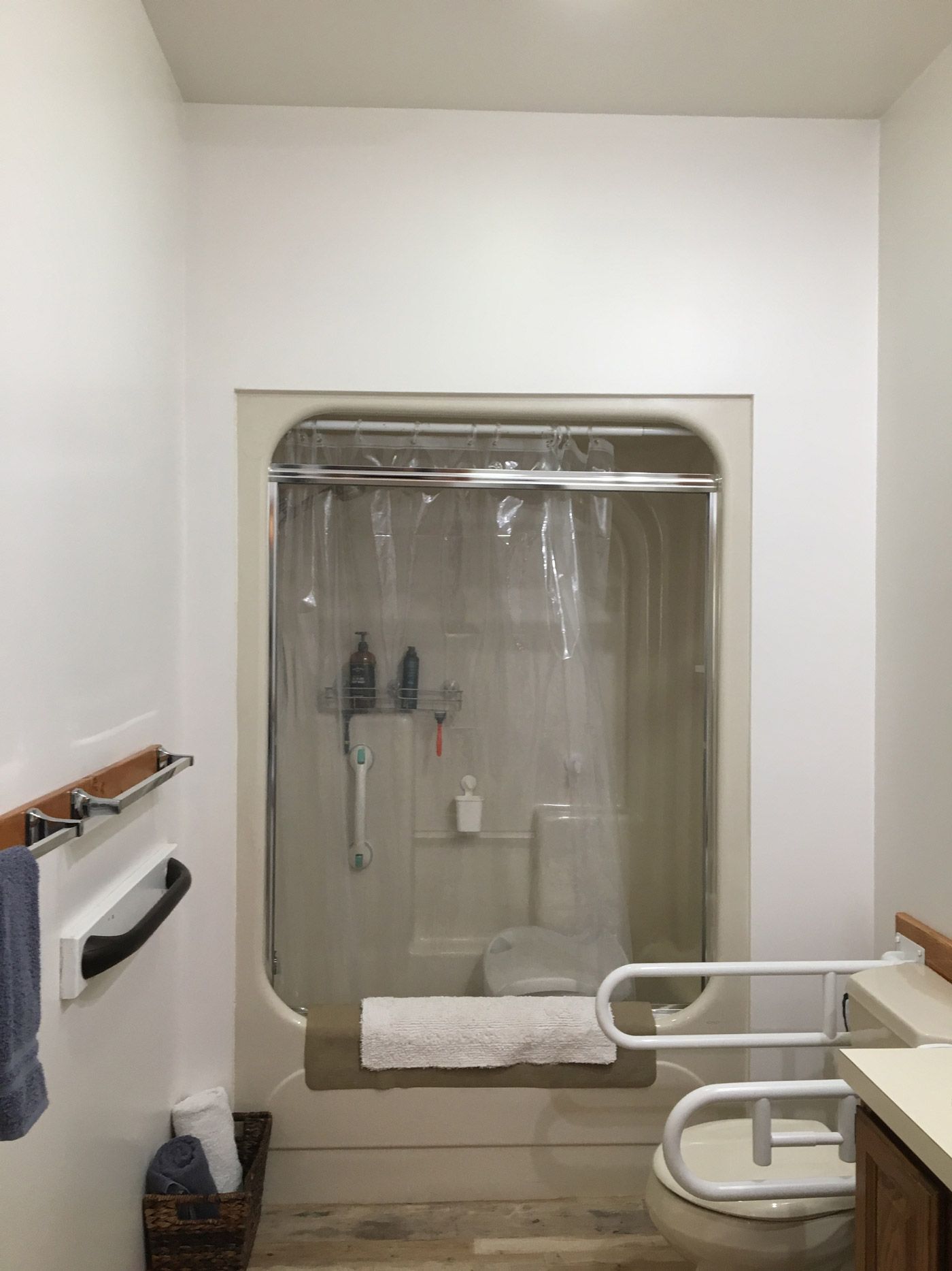 A bathroom features a shower stall with a clear curtain, a toilet with a white safety grab bar, and light-colored walls.