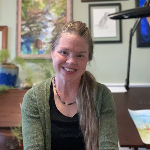 Woman smiling in a green cardigan, seated in an office with framed artwork and a plant in the background