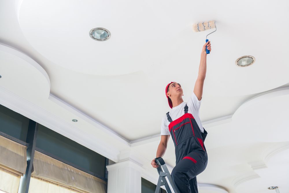 A man is standing on a ladder painting the ceiling with a paint roller.