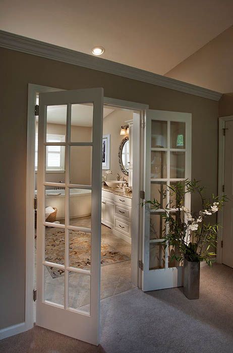 Open white French doors leading to a light-filled bathroom. Beige walls, a rug, and a plant complete the scene.