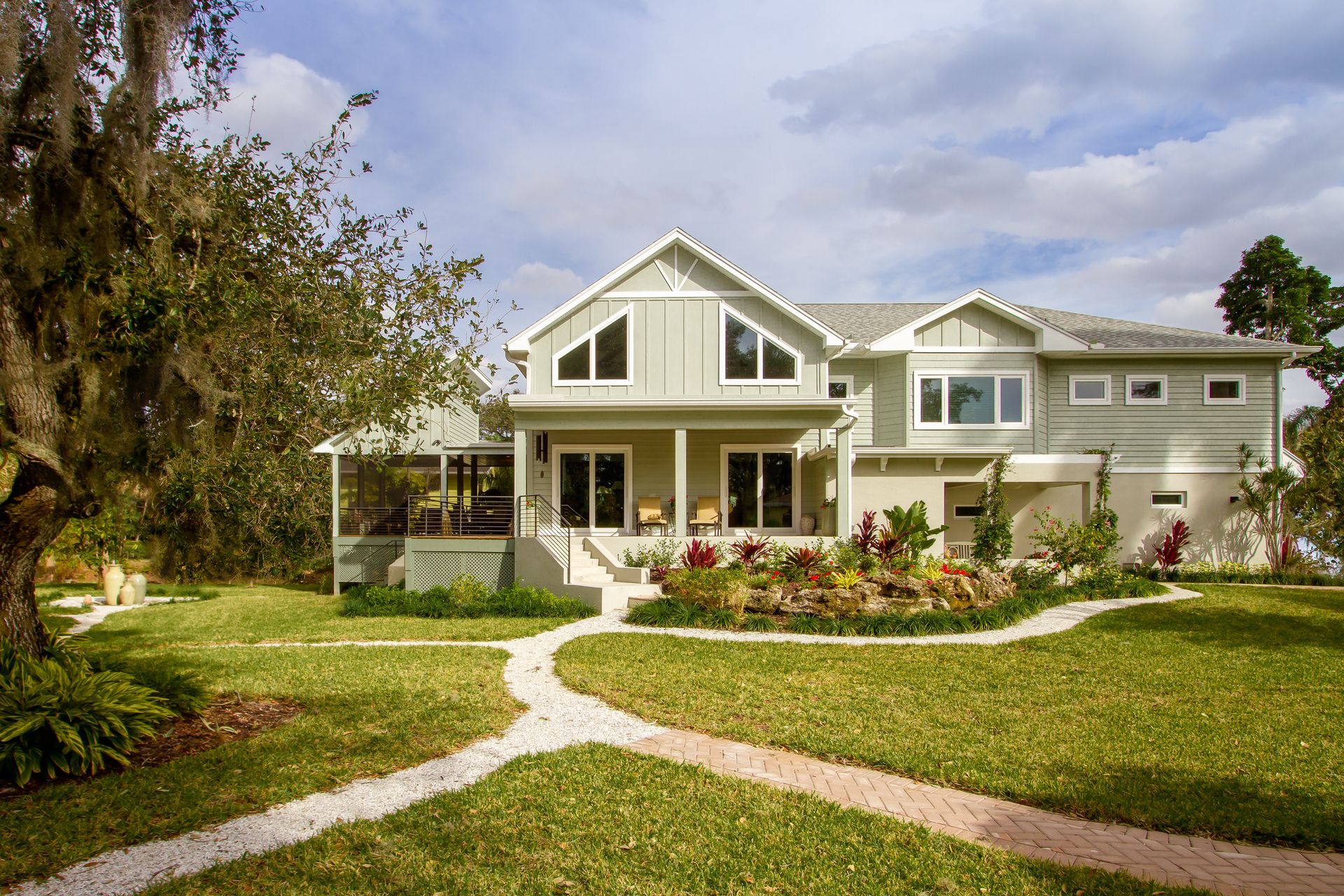 Two-story light green house with gray roof, porch, and a landscaped garden; a pathway leads to the entrance.