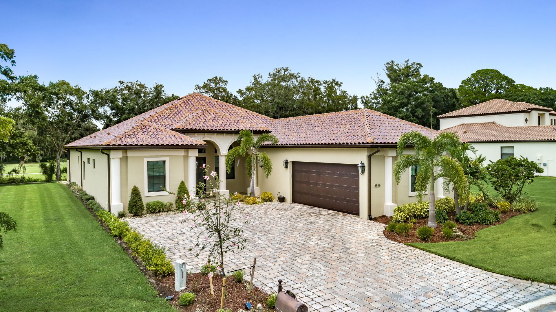 Beige stucco house with a terracotta roof and brick driveway. Palm trees and green lawn.