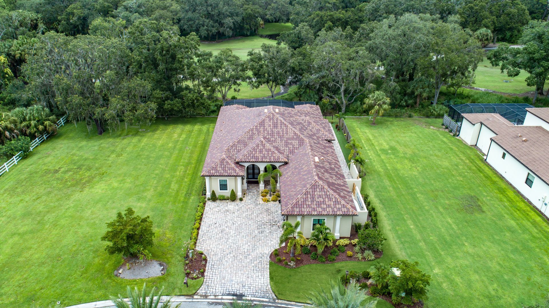 Aerial view of a beige house with a brown tiled roof and a stone driveway on a green lawn surrounded by trees.