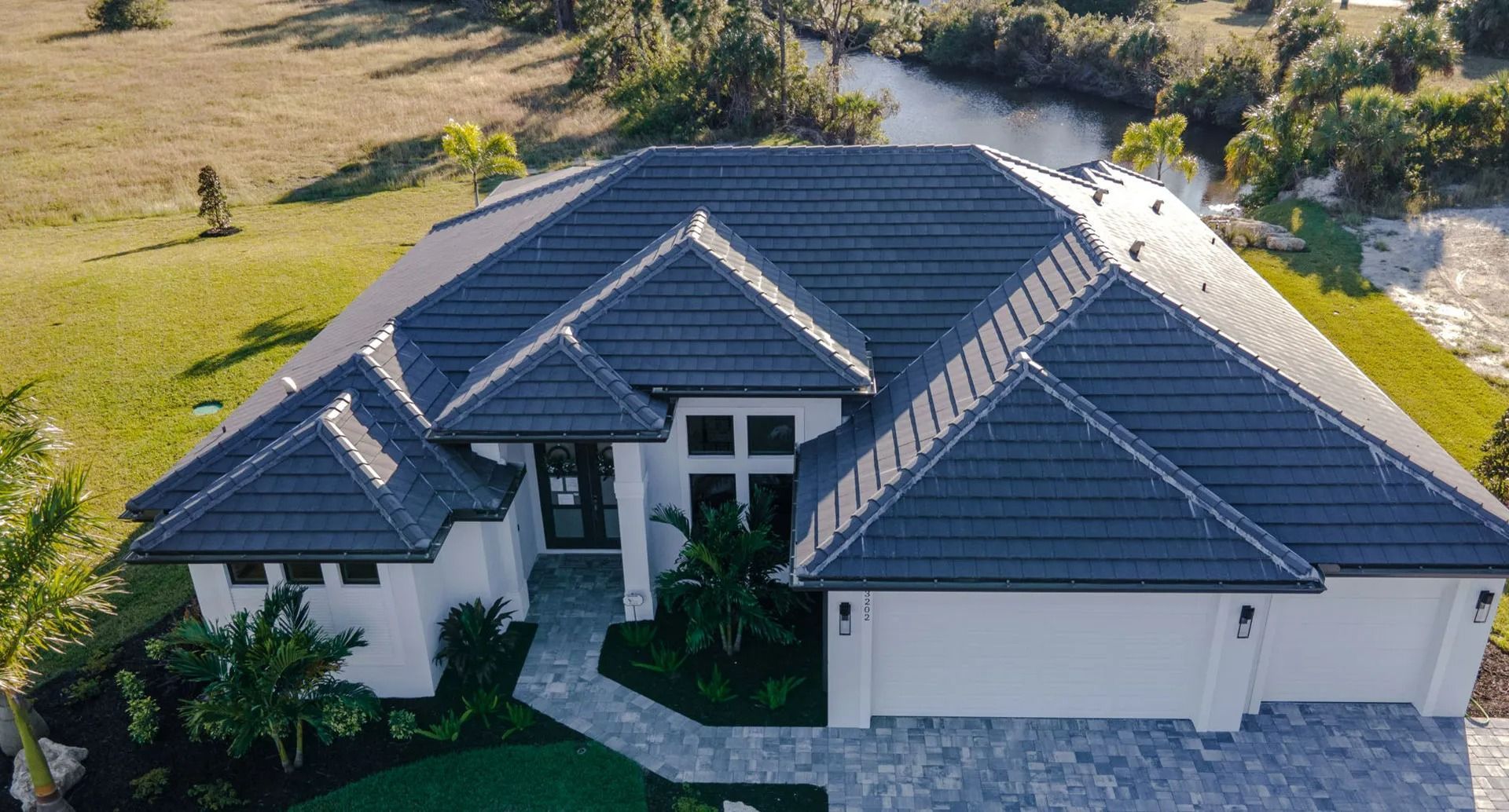 Aerial view of a modern white house with a dark gray tile roof, near a canal and grassy land.