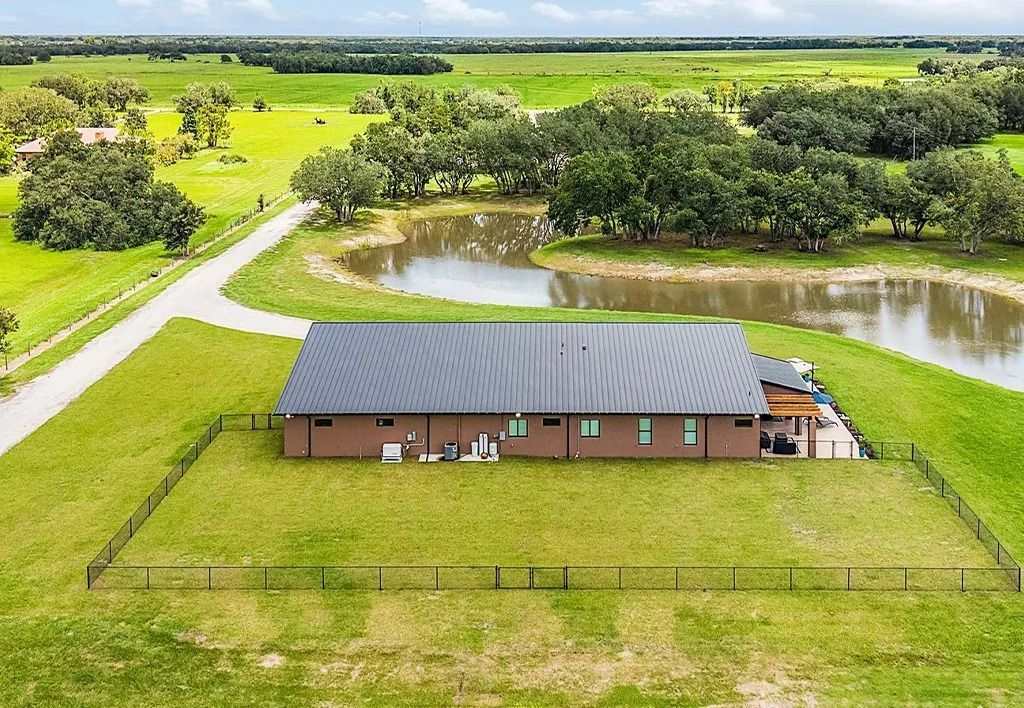 Aerial view of a brown home with a dark roof next to a pond and grassy field.