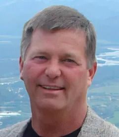 Man smiling, wearing a jacket, with a scenic background of a river and mountains.