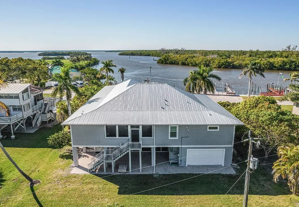 Gray waterfront house with metal roof, palm trees, and water view.