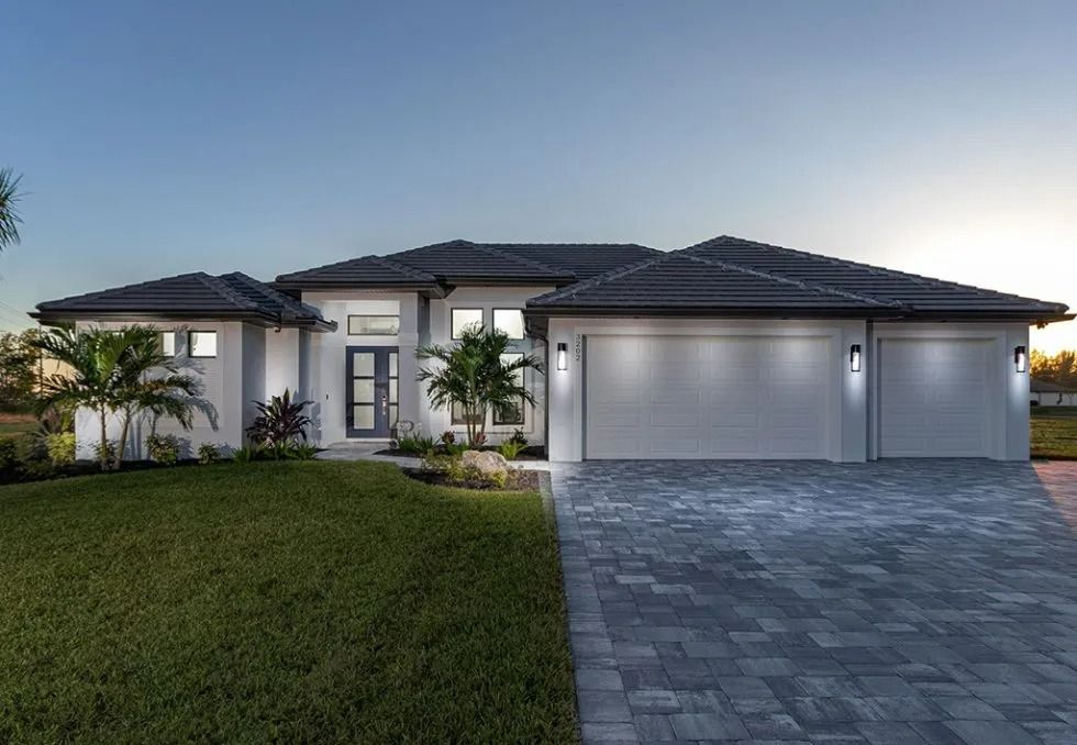 Modern, light-colored house with a three-car garage. Palm trees and green lawn in front, blue sky.