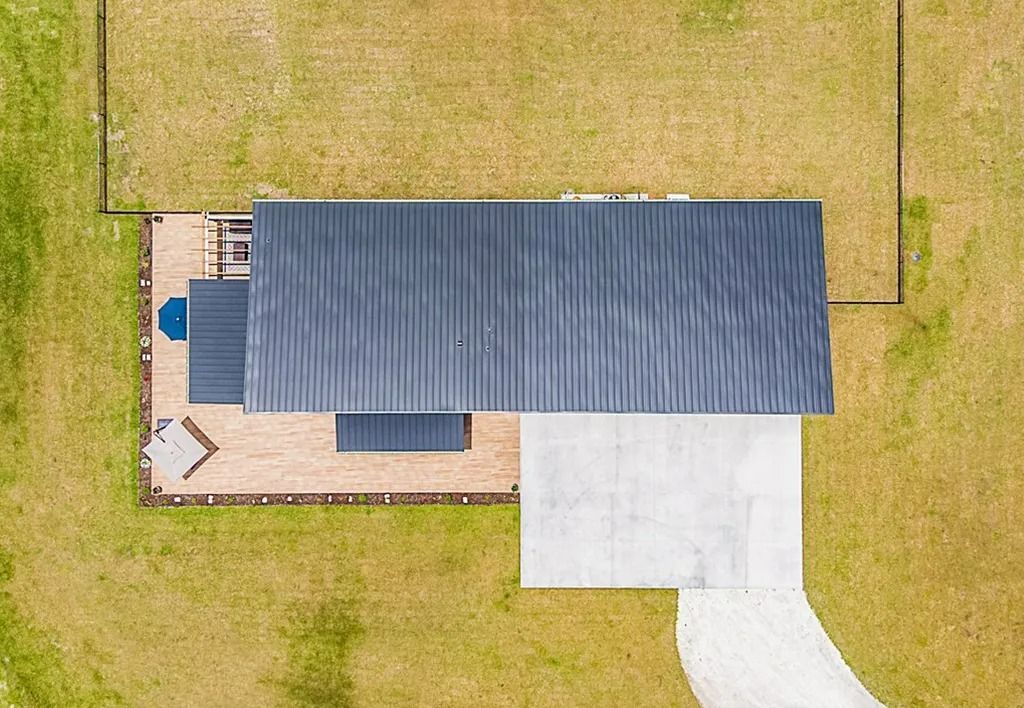 Overhead view of a modern house with a dark metal roof, deck, and concrete driveway on a grassy field.