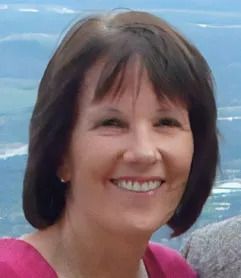 Woman with short dark hair smiles, wearing a pink top, overlooking a landscape.