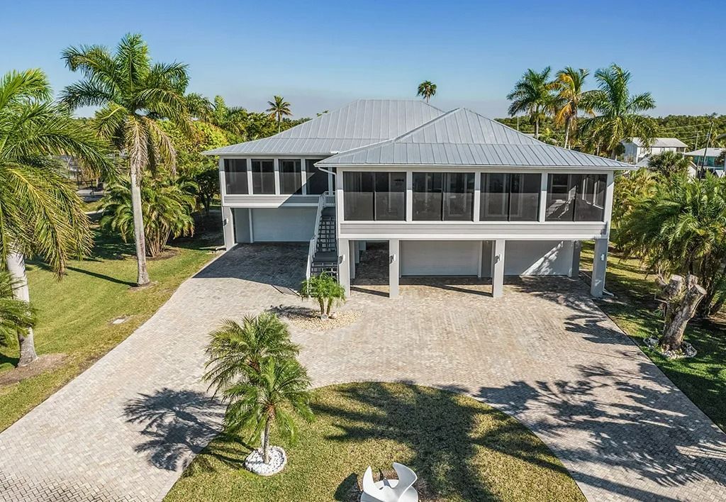 Elevated coastal home with screened porch, surrounded by palm trees and gravel driveway under a blue sky.