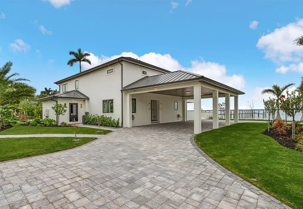 White house with covered parking, brick driveway, and waterfront view under a blue sky.