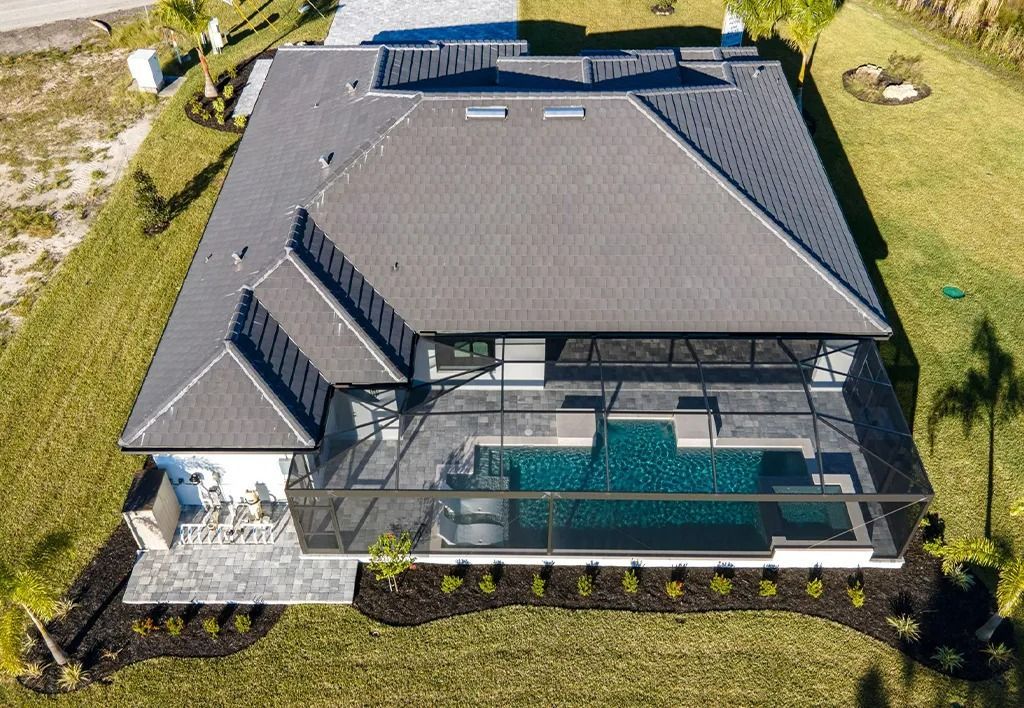 Aerial view of a gray-roofed house with a pool enclosed by a screen. Green lawn and landscaping surround.